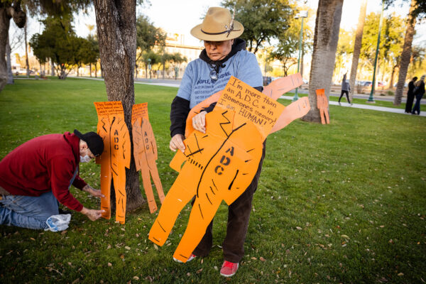 Person wearing a hat holding a cardboard cut-out of an orange Arizona Department of Corrections prison uniform.