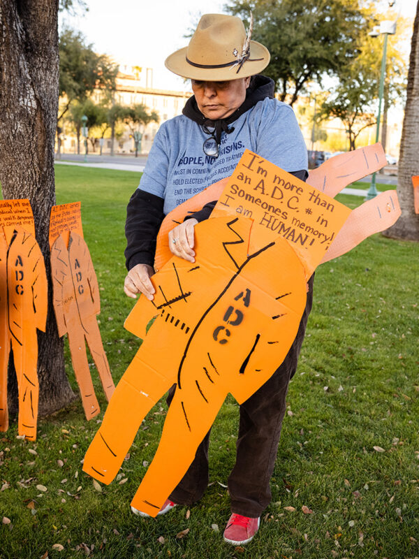 Person wearing a hat holding a cardboard cut-out of an orange Arizona Department of Corrections prison uniform.