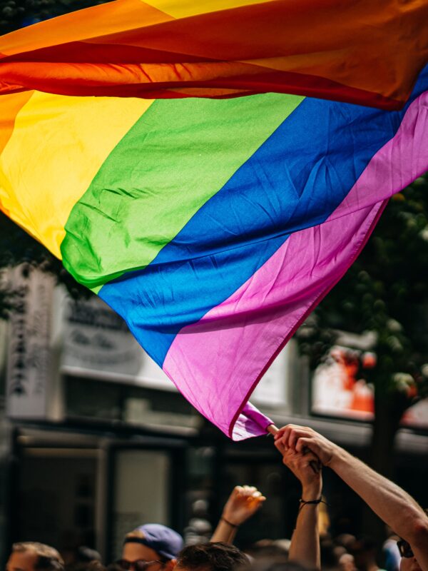 A person's arms waving a Pride flag at a march in a city.