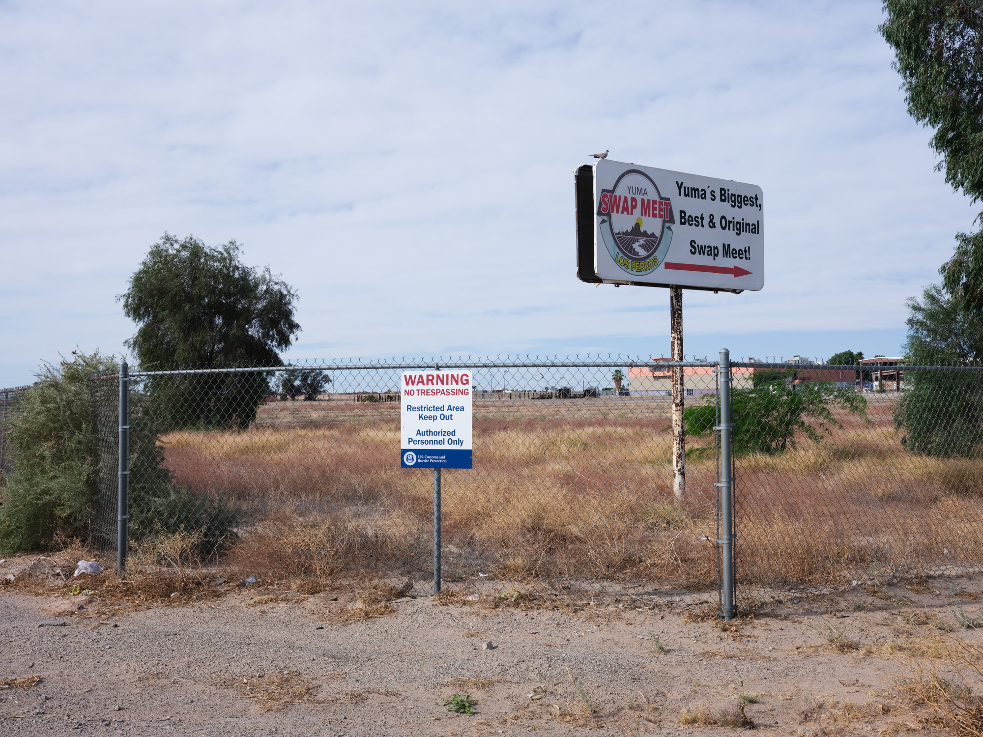 The Yuma County Swap meet billboard sign is seen above a chain-link fence that has a "No trespassing" sign by U.S. Customs and Border Patrol.