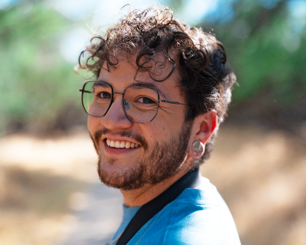 A young trans man with curly black hair, facial hair, and wire framed glasses smiling and wearing a blue t-shirt.