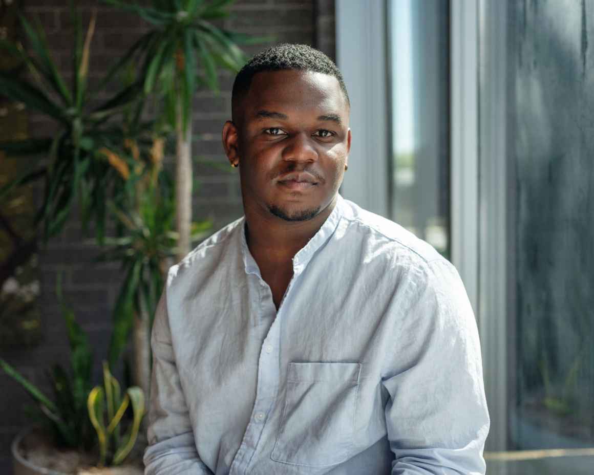 A photo of Malcom, a young Black man with short hair, a small goatee, light blue linen shirt and white pants. His Hands are folded in his lap and has a calm, serious expression on his face. He is seated next to a window in front of an indoor palm tree.