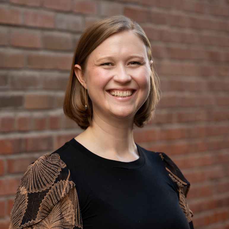 Smiling woman with short, blonde-brown hair standing in front of a brick wall wearing a black blouse.