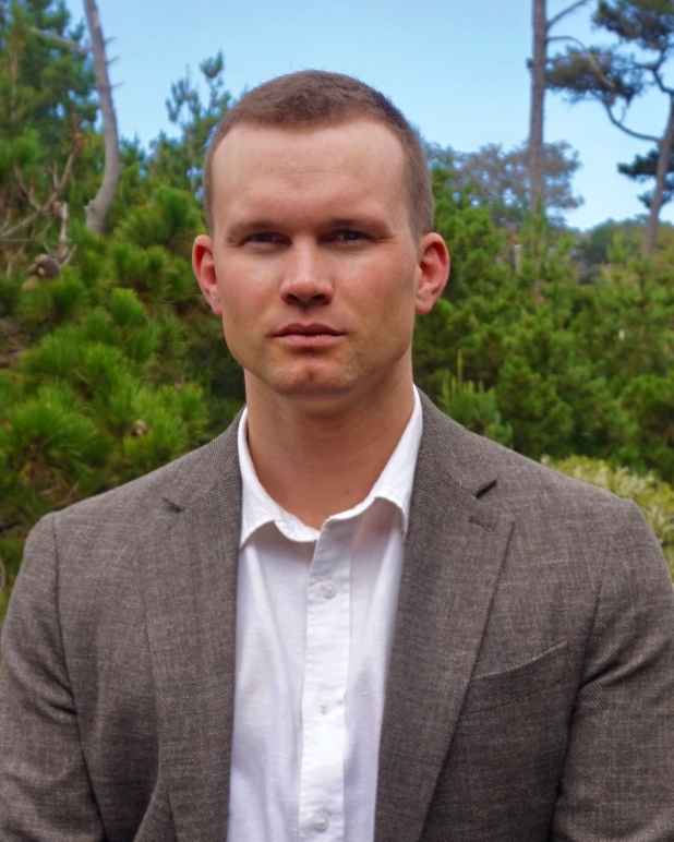 Man with blonde hair in a grey suit with a white shirt in front of a background with trees.