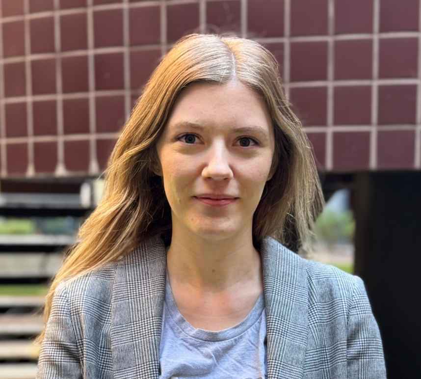 Woman with long blond hair in a grey blazer and blue shirt in front of a red tile background.