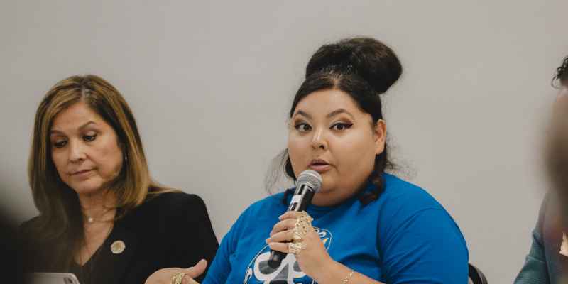 Gina Mendez speaking into a microphone wearing a LUCHA blue shirt.